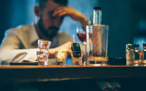 Ein Mann mit Kopfschmerzen sitzt am Tresen, neben Autoschlüsseln in einem Schnapsglas. Alkohol am Steuer unter 21 ist eine Gefahr.