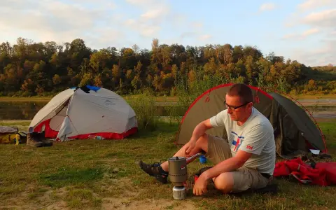 Mężczyzna siedzi przy kuchence turystycznej obok namiotu, przygotowując posiłek. W tle jezioro i las.