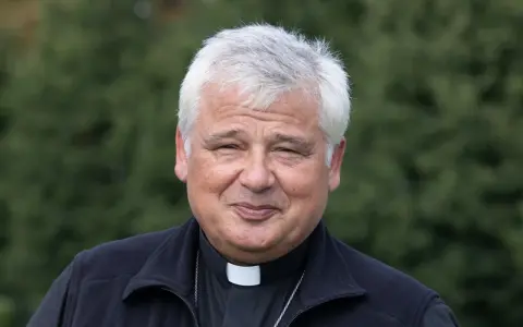 Uśmiechnięty jałmużnik papieski w sutannie i czarnym polarze, na tle zieleni.