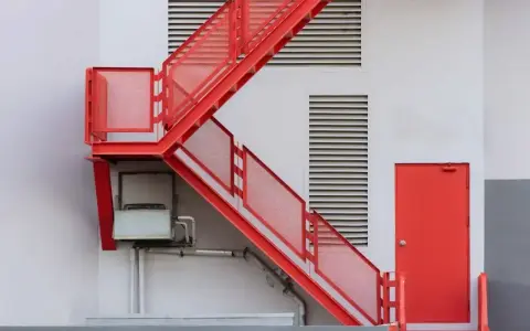 Nowoczesna, czerwona zabudowa schodów zewnętrznych z metalowymi stopniami i ażurowymi barierkami, prowadząca do czerwonych drzwi.