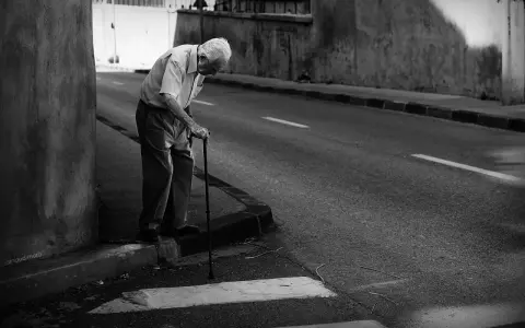 Une photo dans la rue : un vieil homme se penche sur sa canne, le regard perdu, au bord d'une route déserte.