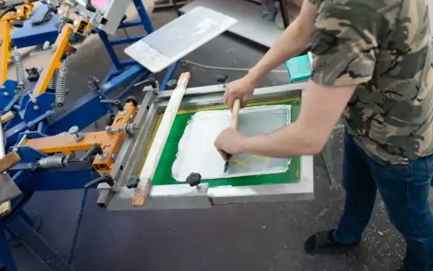 Proceso de serigrafía que es: un operario aplica tinta blanca sobre una pantalla para estampar.