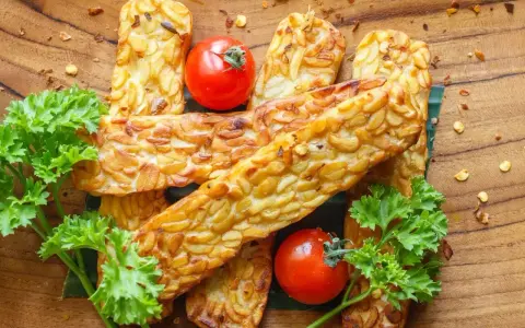 Złocisty, smażony tempeh, czyli co to jest? Podany z pomidorkami i natką pietruszki na drewnianej desce.