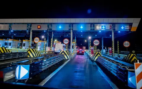 Nocny widok na bramki autostradowe z niebieskimi światłami i znakami, samochody wjeżdżają na płatne pasy.