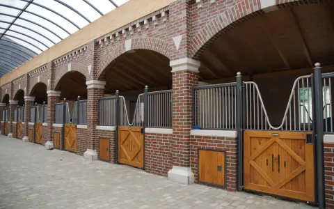 Belle écurie pour chevaux avec des boxes en briques rouges, des portes en bois et un toit transparent.