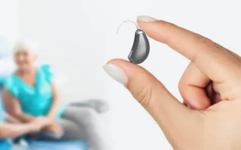 Dłoń trzyma aparat słuchowy. W tle starsza kobieta.