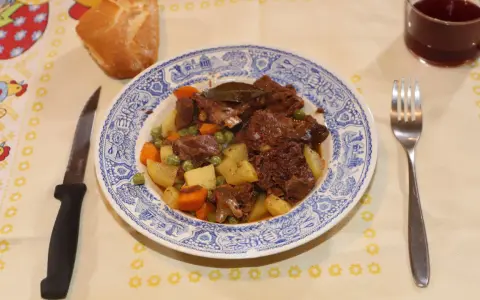 Un plato de corzo guisado con patatas, zanahorias y guisantes, acompañado de pan y vino tinto. ¡Una de las mejores recetas de corzo guisado!