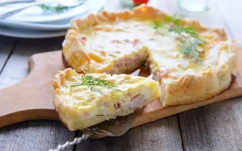 Una fetta di torta salata prosciutto e formaggio, guarnita con aneto, su un tagliere di legno.