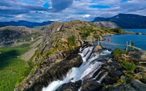 En person på en hängbro över ett vattenfall under en vandringsresa i Norge. Dramatiskt landskap med berg och en blå sjö.