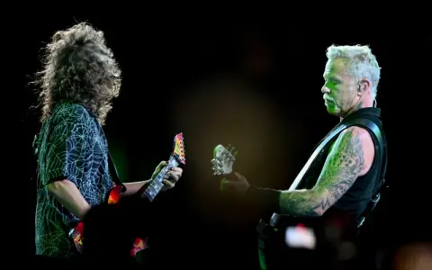 James Hetfield, z charakterystycznym wąsem i wytatuowanymi ramionami, gra na gitarze. Obok niego drugi muzyk z burzą włosów.