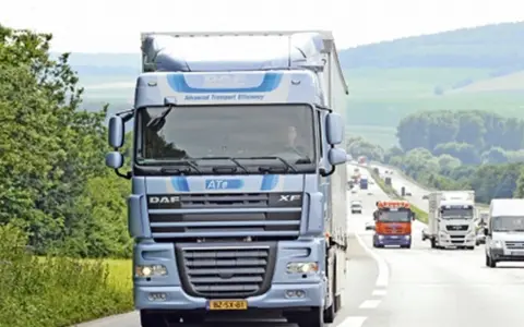 Niebieska ciężarówka DAF XF 105 na autostradzie, otoczona zielenią. W tle inne pojazdy i pagórkowaty krajobraz.