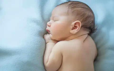 Śpiący skrajny wcześniak na błękitnym kocyku. Delikatne rysy twarzy, małe rączki złożone pod brodą.