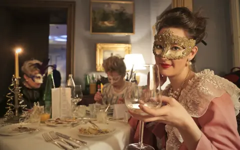 Elegancka kobieta w złotej masce i perłach świętuje karnawał 2026. Wznosi kieliszek wina, otoczona innymi gośćmi w maskach.