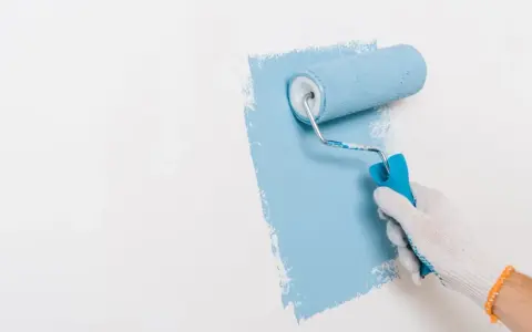 Ręka w białej rękawiczce maluje ścianę na niebiesko wałkiem. Widać nierówności, co może sugerować wady farby ceramicznej.