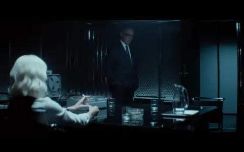 Simon Jacques attore in un ufficio buio, con una donna dai capelli biondi che fuma.
