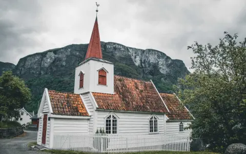 Biały kościółek z czerwoną wieżą i dachem, otoczony białym płotkiem, na tle surowych gór. Symbol norweskiej religii i spokoju.