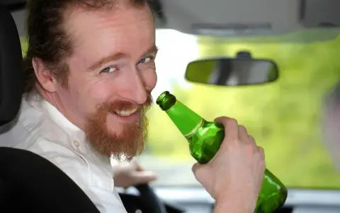 Uśmiechnięty mężczyzna z rudą brodą trzyma zieloną butelkę piwa w samochodzie. Mówi, że piwo to nie alkohol, ale jazda z nim jest niebezpieczna.