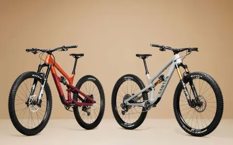 Two full-suspension mountain bikes, one orange and maroon, the other grey, are displayed side-by-side, showcasing the 27.5 vs 29 wheel size debate.