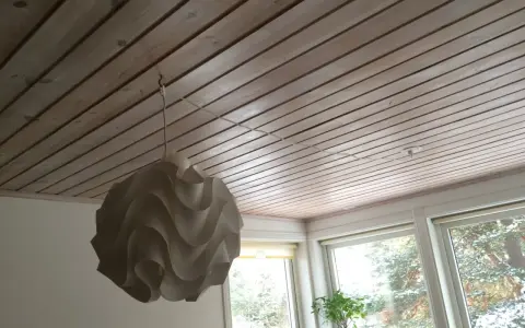 En vit lampskärm hänger från ett tak av träpanel. En tunn sladd dras i taket för att ansluta lampan.