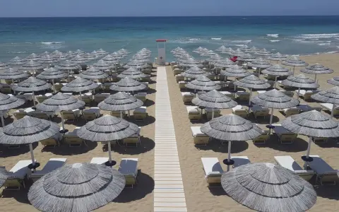 Filettato di ombrelloni di paglia e lettini sulla sabbia dorata, con il mare azzurro sullo sfondo. Un paradiso a Presicce Lido Marini.