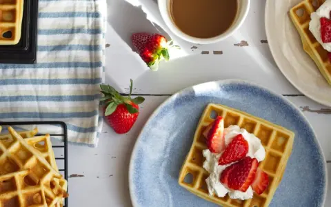 Frische belgische Waffeln mit Erdbeeren und Sahne auf einem blauen Teller, daneben eine Tasse Kaffee und weitere Waffeln auf einem Gitter.