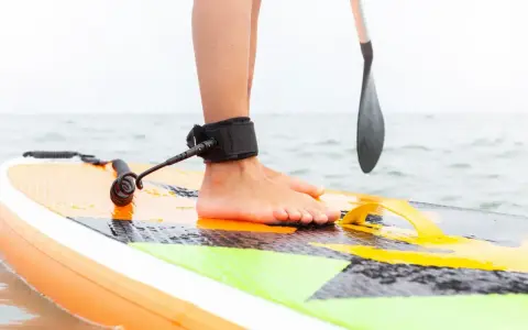 Noga na desce SUP z leash'em, niezbędne akcesoria do sup dla bezpieczeństwa. Wiosło w tle.