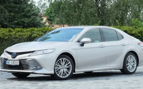 Srebrna Toyota Camry 2.5 Hybrid stoi na wybrukowanym podjeździe, otoczona zielenią.