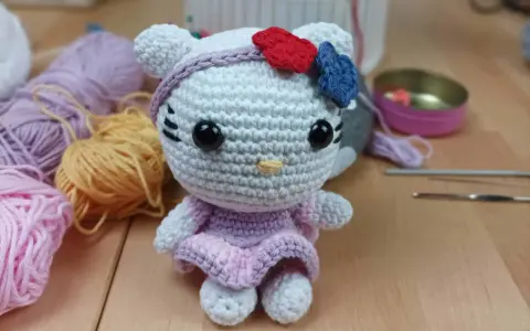 Muñeca de Hello Kitty tejida a crochet, con vestido lila y lazo rojo y azul. Hilos y agujas de crochet alrededor, perfectos para crear tus propios hello kitty crochet patrones español.