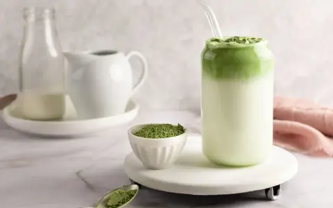 Pyszny matcha latte w szklance, obok miseczka z zielonym proszkiem i łyżeczka.