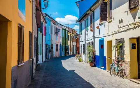 Un vicolo colorato a San Giuliano, con case dipinte di giallo, blu, verde e rosa. Una bicicletta azzurra è parcheggiata davanti a una casa.