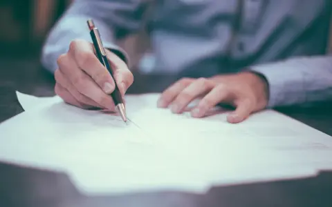 Mano firmando un contrato de reserva de vivienda. El documento se extiende sobre una mesa, con un bolígrafo en acción.