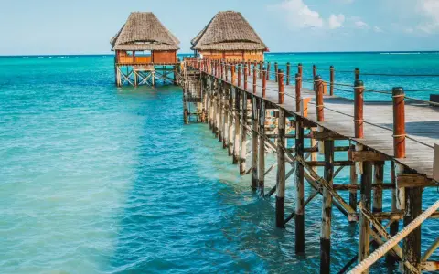 Drewniany pomost prowadzi do chat na palach nad turkusową wodą. Zanzibar, pora deszczowa, ale słońce świeci.