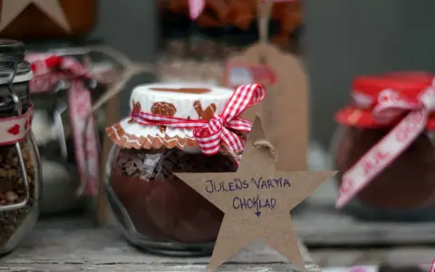 Burkar med ingredienser för varm choklad jul, dekorerade med röda band och pepparkaksfigurer. En stjärna med texten "Julens varma choklad" hänger framför.