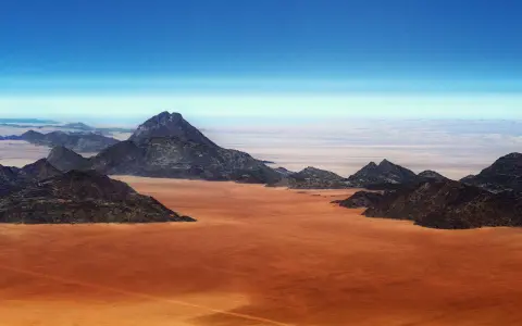 Pustynny krajobraz w **Afryce, Namibia**. Czerwona ziemia i skaliste góry pod błękitnym niebem.
