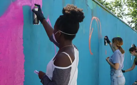 Ungdomar målar graffiti på en blå vägg. En flicka med afrohår sprayar rosa färg.