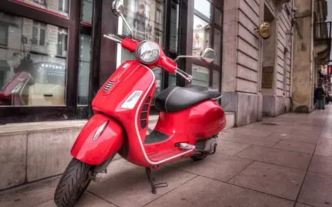 Czerwony skuter Vespa zaparkowany na chodniku przed budynkiem.