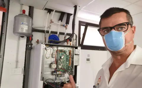 Técnico con mascarilla realiza mantenimiento aerotermia. Equipo abierto, circuito visible. Pulgar arriba.