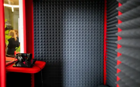 Cabina insonorizzata con pannelli fonoassorbenti che assorbono il suono. Cuffie e tablet su un ripiano rosso.