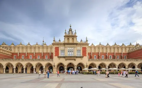 Sukiennice w Krakowie, zabytkowy budynek z arkadami, tętni życiem. Ludzie spacerują po Rynku Głównym, podziwiając architekturę.