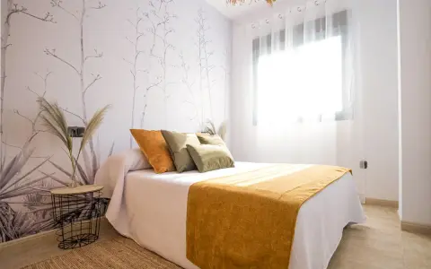 Habitación con papel tapiz botánico y cama con cojines naranjas y verdes. Una manta mostaza ilumina el espacio, ideal para quienes buscan colores para paredes con gotele.