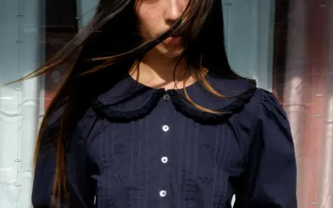A young woman with windswept hair wears a dark blue blouse with a charming peter pan neckline, delicate embroidery, and puffed sleeves.