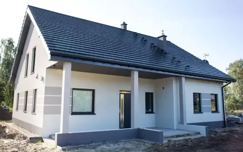 Nowy dom z szarą dachówką i jasną elewacją, ozdobioną subtelnymi, poziomymi pasami. Trwają prace nad malowaniem elewacji, nadając budynkowi nowoczesny wygląd.