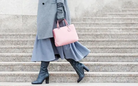 Stilvoll **Kleider im Winter tragen**: Grauer Mantel, blauer Rock, schwarze Stiefel und eine rosafarbene Tasche auf Steinstufen.