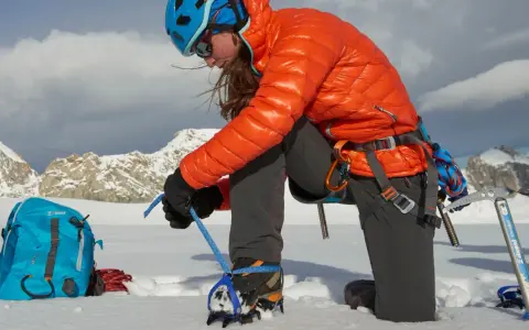 Alpinistka zakłada raki górskie na buty, przygotowując się do wspinaczki w śnieżnych górach.