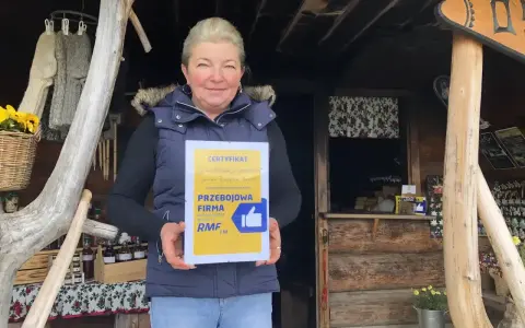 Kobieta z certyfikatem "Przejściowa Firma" przed bacówką. W tle widać rękodzieło i przetwory, a także tablicę z napisem "Bacówka u Jancora".