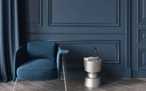 Elegante sillón azul junto a una mesa auxiliar plateada, con cortinas a juego y detalladas molduras en paredes de tono oscuro.