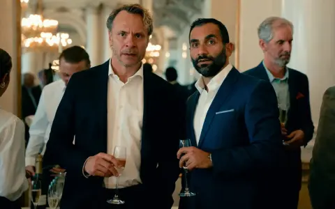 Två män i kostym håller glas med champagne. Bakom dem syns en festlig miljö med ljuskronor. Kanske en scen från "hundarna" på SVT.