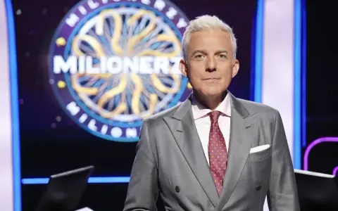 Hubert Urbański w garniturze, na tle koła z napisem "Milionerzy". Co dajemy komuś na drogę milionerzy? Wiedzę i odwagę!