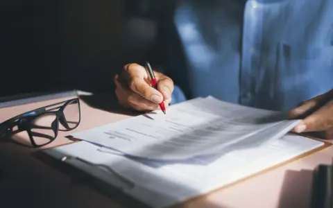 Persona revisa y firma una nota de encargo inmobiliaria, con gafas y bolígrafo rojo sobre documentos.