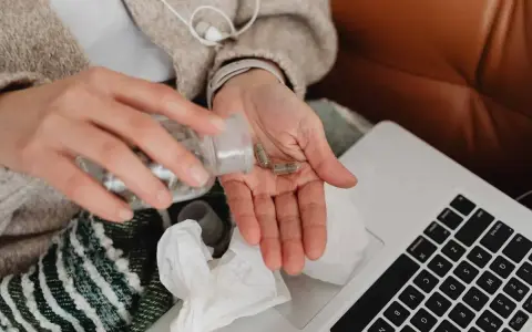 Dłoń z kapsułkami leków, obok laptop i chusteczki. Kto płaci za L4, gdy choroba dopada w domu?